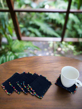 Load image into Gallery viewer, Set of black coasters with colorful edges on a wooden table, one coaster with a white mug.