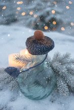 Load image into Gallery viewer, A handmade crocheted mushroom cap with a brown stem and pom-pom sits on top of a clear glass bottle, set against a tiled floor background.