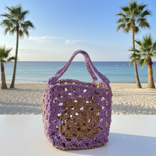 Load image into Gallery viewer, A handmade crochet bag in purple and brown yarn, featuring a large central crochet motif and a top handle, sitting on a white table with a wall, palm trees, and blue sky in the background.