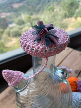 Load image into Gallery viewer, A pink crocheted hat with a flower accent and a small dangling tail, displayed on a clear glass bottle in front of a window with a blurred outdoor landscape.