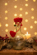 Load image into Gallery viewer, A clear glass bottle with a bulbous base, decorated with a bright red crocheted hat and matching red cord accessories, photographed outdoors against a white wall and mulch background.
