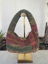 Load image into Gallery viewer, A handmade crocheted shoulder bag with a large arching handle, displayed on a gold stand. The bag features textured stripes of olive green, teal, orange, pink, and gold yarn, with a matching tassel detail on the side.