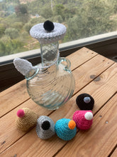 Load image into Gallery viewer, A clear glass bottle with a crocheted white neck cover and black pom-pom lid sits on a wooden table, with five small crocheted owl-themed ornaments arranged in front. A window with greenery outside is visible in the background.