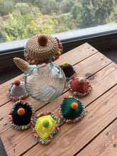 Load image into Gallery viewer, A set of crocheted teapot and cup lids with colorful pom-pom trim, arranged on a wooden table in front of a window showing a green outdoor view.