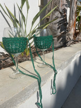 Load image into Gallery viewer, Two clear wine glasses wrapped in dark green crocheted mesh bags with long dangling straps, placed on a concrete ledge outdoors with plants and a wall behind them.