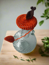 Load image into Gallery viewer, Glass pitcher with a crocheted orange lid and brown handle on a wooden surface.