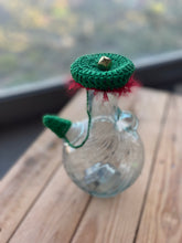 Load image into Gallery viewer, Decorative glass bottle with green crochet top and red fabric, on a wooden surface.