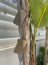 Load image into Gallery viewer, Two small woven shoulder bags in a taupe-beige color hang from a dried palm tree trunk, with a white shuttered wall and a green banana leaf in the background.