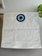Load image into Gallery viewer, White towel with a blue and black evil eye design on a beige background