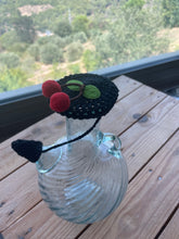 Load image into Gallery viewer, A clear ribbed glass bottle or vase with a black crocheted cherry-themed lid or cover, featuring two red felt cherries and green felt leaves, displayed on a wooden table with a blurred outdoor landscape in the background.