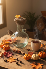 Load image into Gallery viewer, A brown crocheted acorn-shaped hat with a yellow leaf accent and a matching brown tail, displayed on a clear glass bottle against a light gray tiled background.