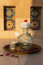 Load image into Gallery viewer, A white crocheted hat with a red flower accent on top, displayed on a clear glass bottle against a light tiled background.