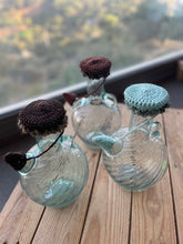 Load image into Gallery viewer, Three glass bottles with knitted covers on a wooden surface with a blurred natural background