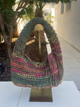 Load image into Gallery viewer, A crocheted shoulder bag with multicolor stripes of green, pink, orange, and yellow, displayed on a gold-colored stand in an outdoor patio setting.