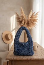 Load image into Gallery viewer, A handmade blue crocheted shoulder bag with a thick textured handle, resting on a light beige fabric surface against a white wall.
