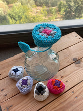 Load image into Gallery viewer, A clear glass bottle with a turquoise crocheted lid and matching handle, displayed on a wooden table with four small crocheted heart-shaped accessories nearby. A window with blurred greenery is visible in the background