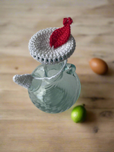 Load image into Gallery viewer, Glass carafe with a crocheted red and white lid on a wooden surface.