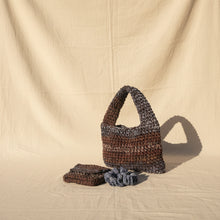 Load image into Gallery viewer, A handmade crocheted tote bag in brown and navy variegated yarn, shown with a matching crocheted pouch and scrunchie on a stone patio next to a tree trunk and green plants.