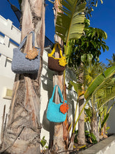 Load image into Gallery viewer, Three knitted bags in blue, brown, and gray on a palm tree branch with a blue sky background.
