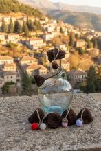 Load image into Gallery viewer, A clear glass bottle with a long neck and a brown crocheted cap, decorated with several small colorful pom-poms on the side, photographed against a light gray concrete wall.
