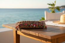 Load image into Gallery viewer, A tan macrame clutch bag with fringe and colorful wooden beads, resting on a stone ledge outdoors with green leaves and a white wall in the background.
