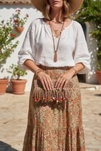 Load image into Gallery viewer, A tan woven fringe clutch bag with colorful wooden beads hanging from the bottom, displayed outdoors on a stone ledge in front of green foliage.
