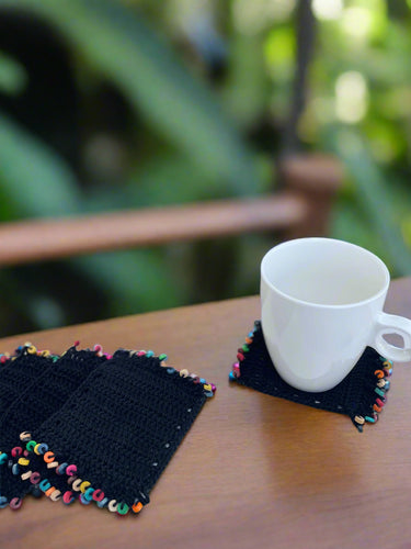 White mug on a black crocheted coaster with colorful tassels on a white surface outdoors.