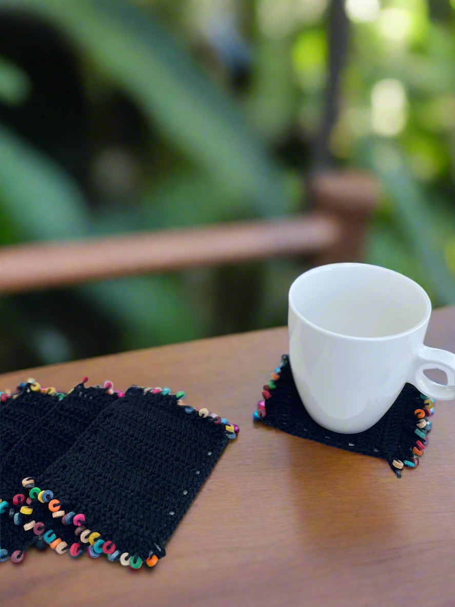 White mug on a black crocheted coaster with colorful tassels on a white surface outdoors.