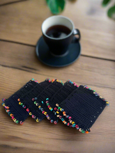 Load image into Gallery viewer, Set of black coasters with colorful pom-poms on a wooden surface next to a cup of coffee.
