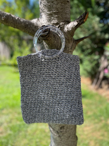 Textured gray bag with a clear plastic handle hanging on a tree branch outdoors.