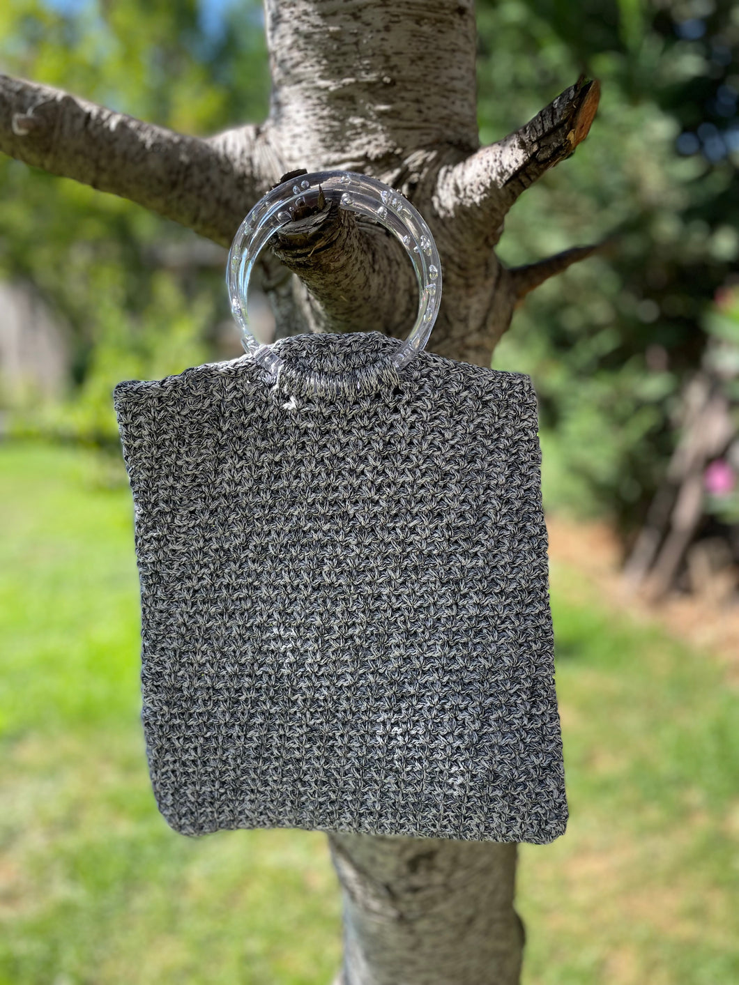 Textured gray bag with a clear plastic handle hanging on a tree branch outdoors.