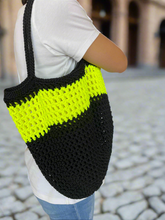 Load image into Gallery viewer, Person holding a black and neon yellow crochet bag on a cobblestone street.
