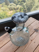 Load image into Gallery viewer, A clear glass bottle with a textured gray and white crocheted cap and a small black-and-white knotted cord, placed on a wooden deck with a blurred outdoor garden background.
