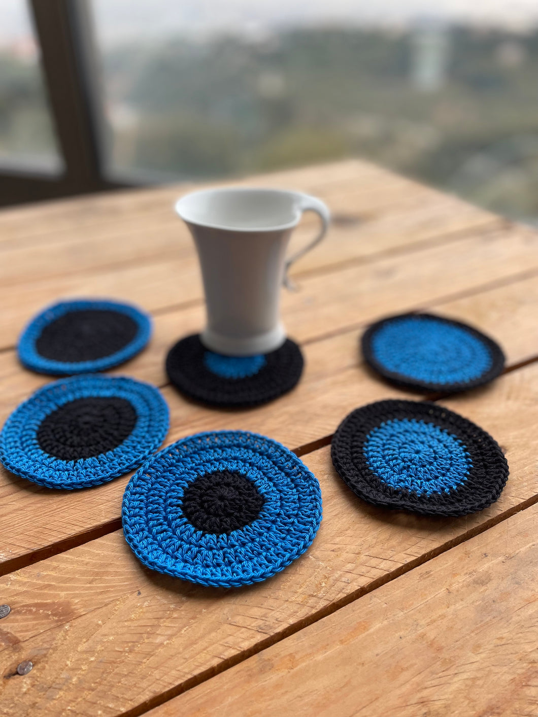 Set of blue and black crocheted coasters with a white mug on a wooden surface.