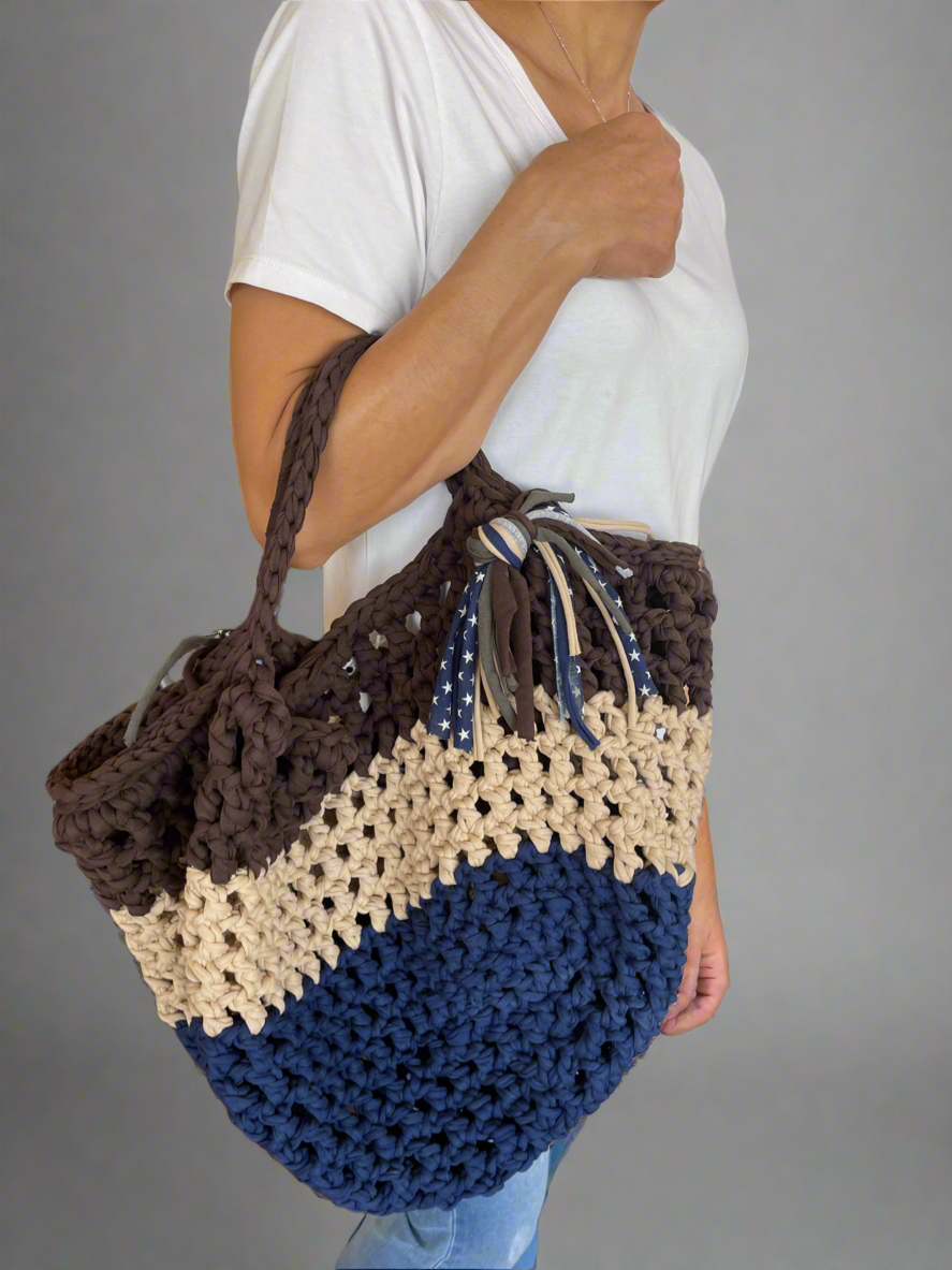 Person holding a crocheted bag with brown, beige, and blue sections against a gray background