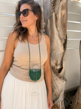 Load image into Gallery viewer, A woman wearing a beige tank top and white skirt models a green crochet wine glass necklace with a long green cord, holding a wine glass to demonstrate its use.
