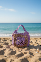 Load image into Gallery viewer, A handmade crocheted tote bag with a purple and brown color-blocked design, featuring a large purple floral motif on the front and dual top handles, set outdoors on a white surface.
