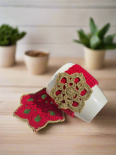 Load image into Gallery viewer, Crocheted red and gold coasters on a white surface with plants in the background
