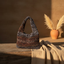 Load image into Gallery viewer, A crocheted shoulder bag in a variegated brown, black, and gray yarn pattern, hanging from a dried palm tree trunk in an outdoor garden setting.
