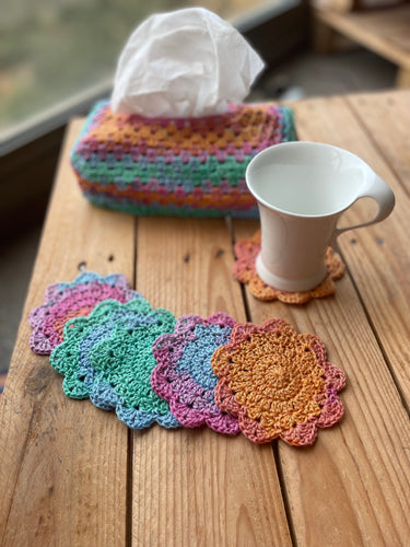 Colorful crocheted coasters on a wooden surface with a tissue box and mug.