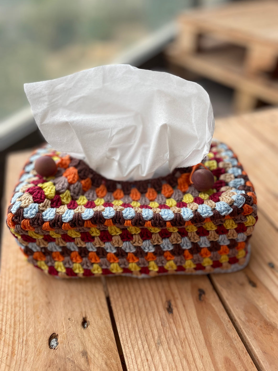 Colorful crocheted tissue box cover with a tissue on a wooden surface