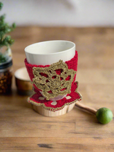 Load image into Gallery viewer, White mug with a red and gold crochet cozy on a wooden surface
