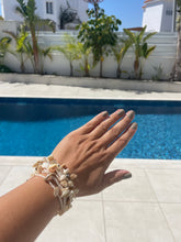 Load image into Gallery viewer, A close-up of a person's hand and wrist wearing a stack of natural beige seashell and pearl bracelets, set against a sunny outdoor background with a swimming pool and white stucco walls.
