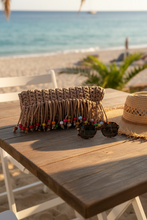 Load image into Gallery viewer, A chunky crocheted clutch bag in natural tan yarn with long fringe and colorful wooden beads, resting on a light-colored stone ledge against a white wall.
