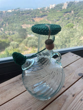 Load image into Gallery viewer, A clear glass bottle with a rounded body, small side handles, and a dark green crocheted stopper on the neck, placed on a wooden table with a scenic outdoor landscape in the background.
