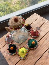 Load image into Gallery viewer, A set of colorful crocheted teapot covers arranged around a clear glass teapot on a wooden table, with a window and greenery visible in the background.
