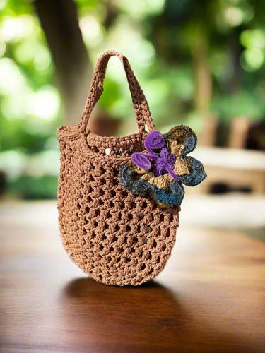 Brown crochet bag with a crochet butterfly charm decoration on a rocky surface.