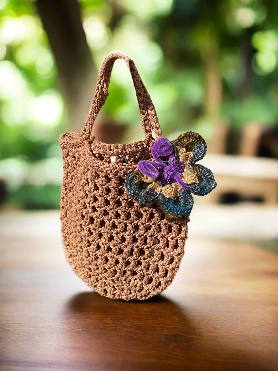 Brown crochet bag with a crochet butterfly charm decoration on a rocky surface.
