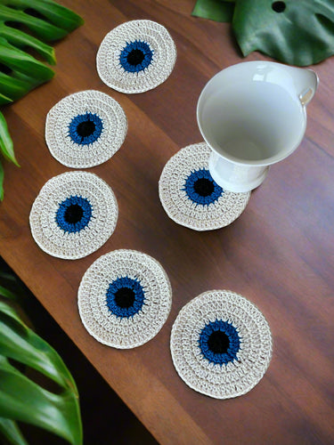 Set of six crocheted coasters with blue centers or evil eye on a wooden surface, one coaster with a white mug.