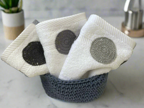 Three white washcloths with black and gray scrubs in a blue basket on a tiled wall background